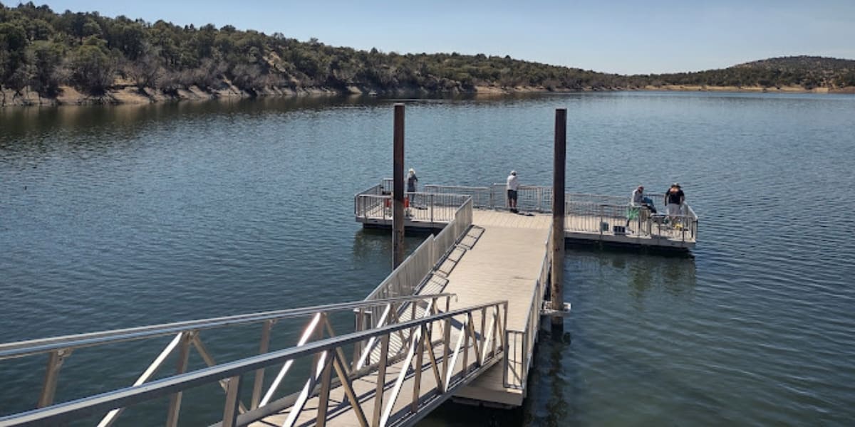 Floating Fishing Platforms connected by wooden walkway extending into a calm lake, with several anglers fishing from the railed platforms while surrounded by tree-covered hills under a clear blue sky.
