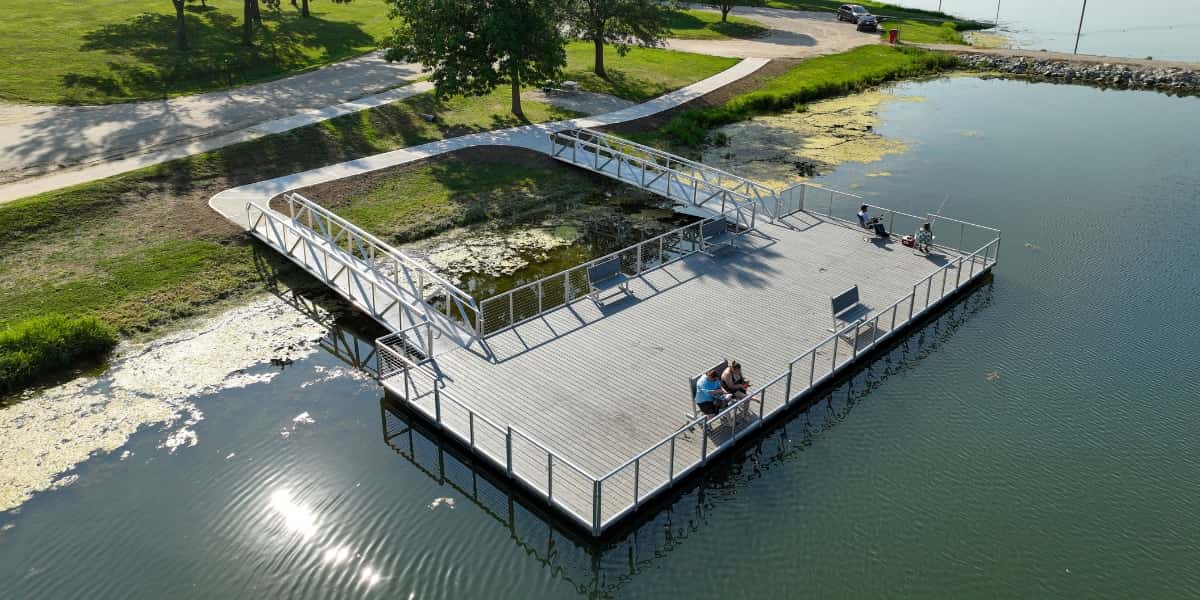 Aerial view of a large fishing platform connected to shore by a metal walkway, demonstrating the pier vs dock design differences, with anglers fishing from the railed platform overlooking a calm lake surrounded by green parkland.