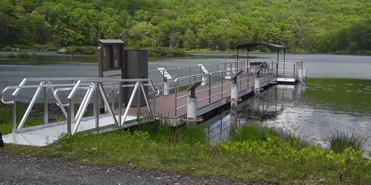 Floating piers with brown decking and metal railings extending into a calm lake, featuring a small utility building and walkway access from a grassy shoreline surrounded by dense green forest.